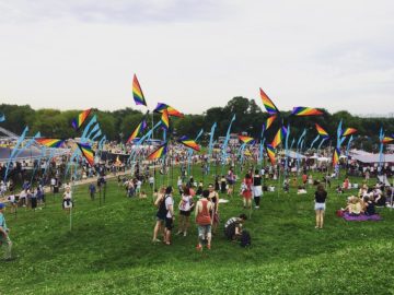 Посетители фестиваля стоят на поляне на фоне ветровых турбин радужного цвета и голубых флагов