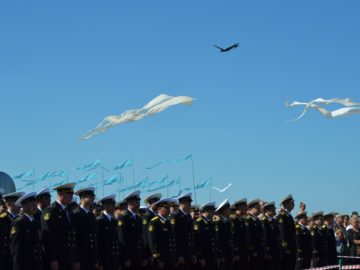 На фоне строя моряков в порадно форме летают белые воздушные змеи и развеваются голубые флаги